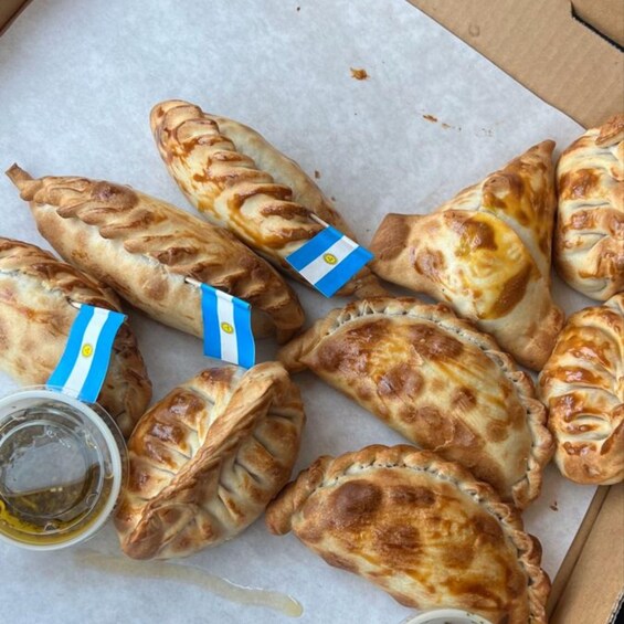 A box of empanadas with a blue and white flag, accompanied by a drink and a sauce container. 3