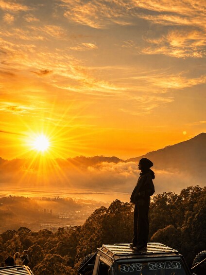 A person standing on a vehicle with a view of a sunset over a mountainous landscape. 4