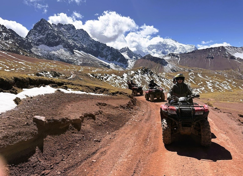 A group of people on ATVs on a dirt road with snow patches and mountains in the background. 1