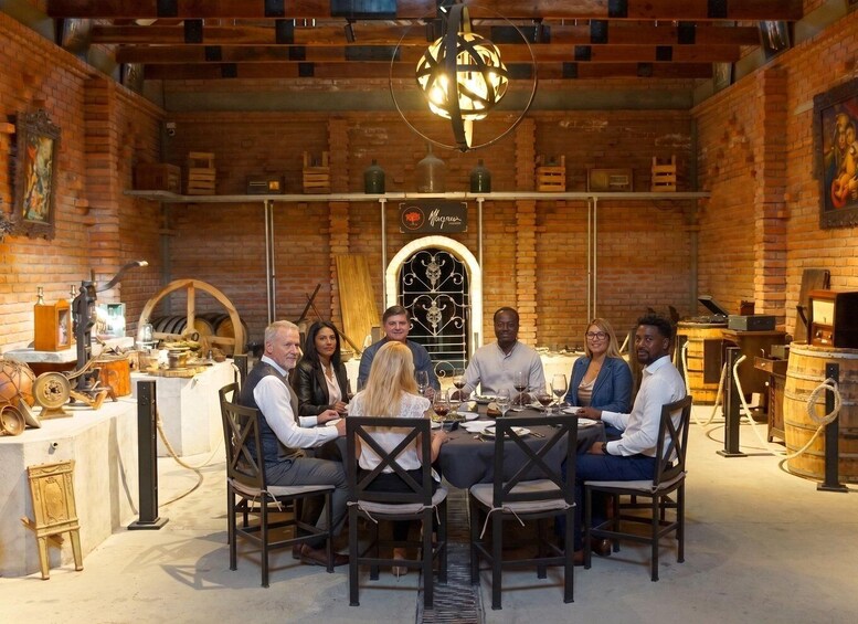 A group of people seated around a table in a rustic brick room with a chandelier, wooden barrels, and vintage equipment. 4