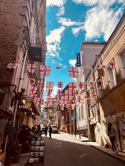 A narrow street with buildings on either side, a clear blue sky, and flags hanging across the street. 2