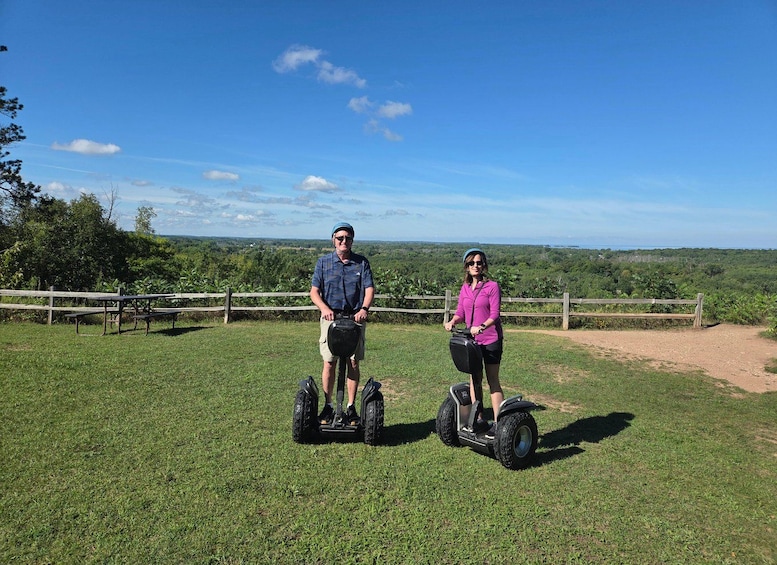 Two people on Segways in a grassy area with a wooden fence and a scenic view of the landscape. 1