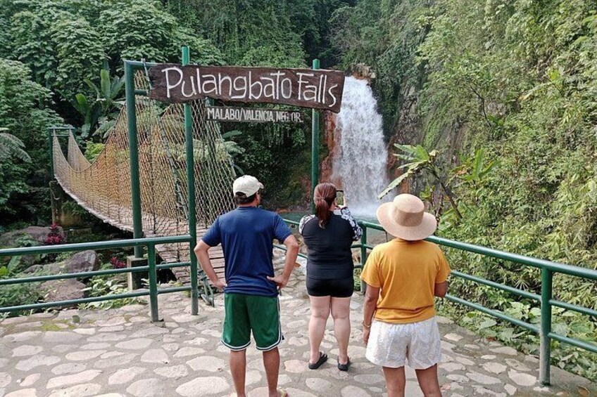 A sign for Pulangbato Falls, a natural waterfall, with people observing it. 2