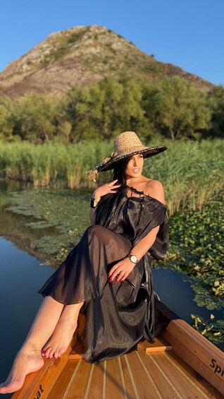 A person sitting on a wooden boat in a calm body of water with greenery and a hill in the background. 1