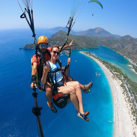 A person and a guide in a harness, paragliding over a coastal area with a beach and mountains in the background. 5