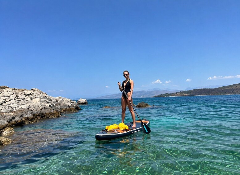 A person paddleboarding on clear turquoise water with rocky shores and distant mountains. 2
