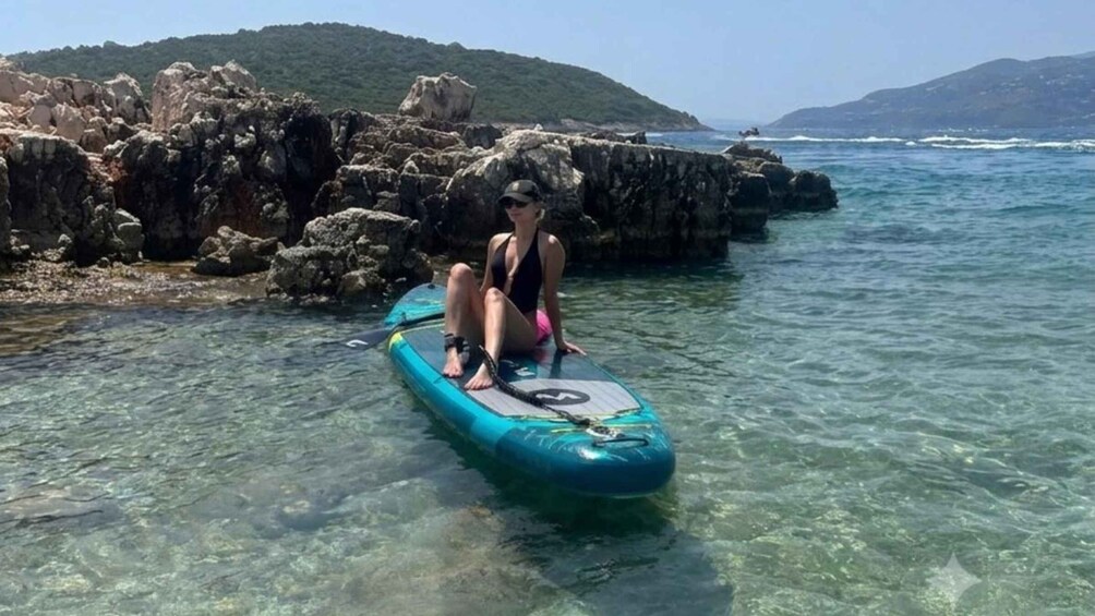 A person sitting on a blue inflatable raft in clear shallow water with rocky terrain and hills in the background. 1