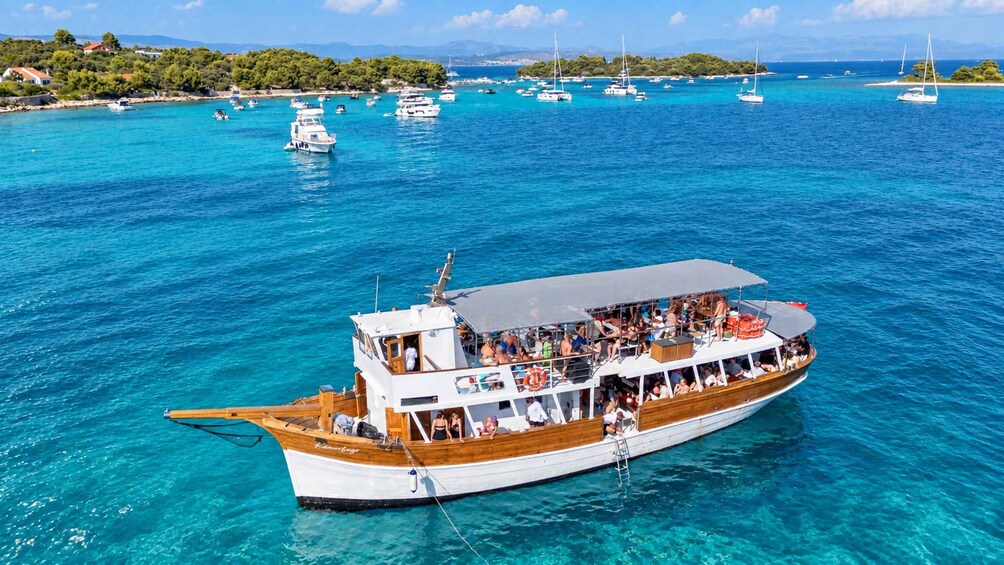 A boat with passengers on a clear blue sea, surrounded by other boats and a coastline with trees and buildings. 1