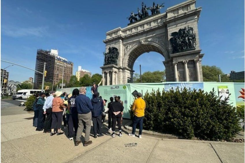 A group of people gathered in front of a large archway with statues on top, standing on a sidewalk with buildings in the background. 5