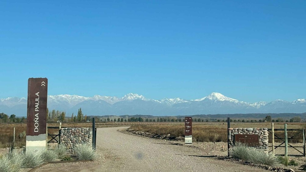 A gravel road leading to a gate with a sign reading 'DONA PAULA' and snow-capped mountains in the background. 2