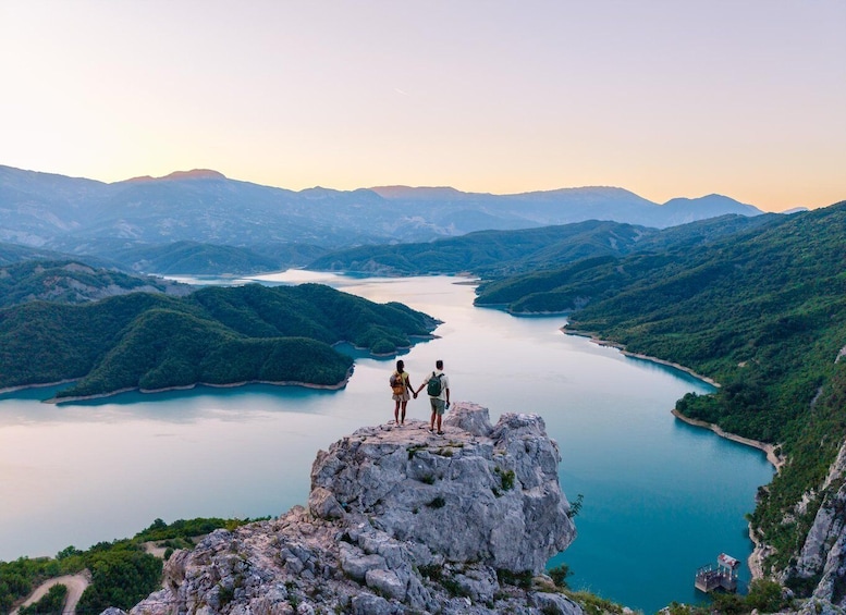 Two people standing on a rocky outcrop overlooking a lake surrounded by mountains. 2