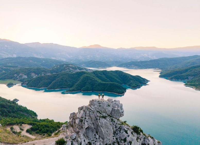A mountainous landscape with a lake and two hikers on a rocky outcrop. 5
