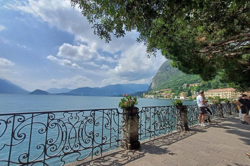 A lakeside promenade with ornate metal railings, potted plants, and a view of distant mountains and buildings. 2