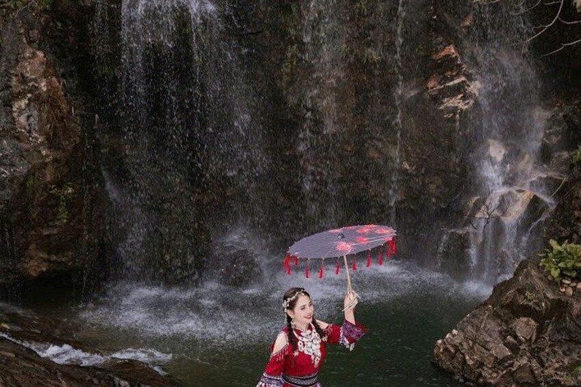 A person in traditional attire holding a red umbrella in front of a waterfall. 4