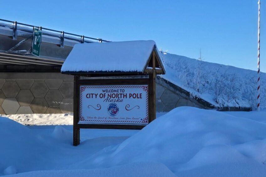 A sign welcoming visitors to the City of North Pole, Alaska, with a snow-covered roof and a clear blue sky in the background. 5