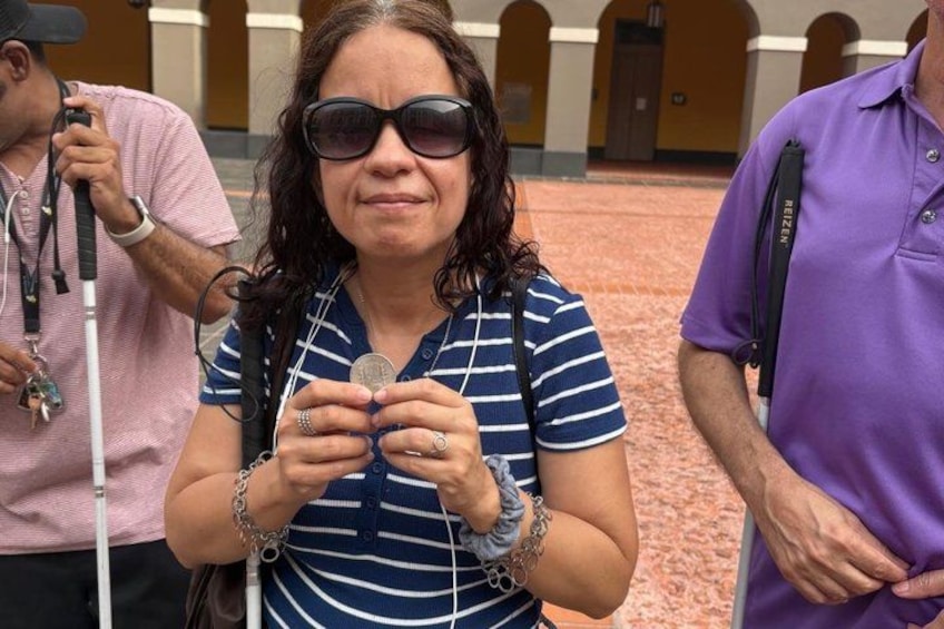 A person holding a coin, standing in an outdoor area with a tiled floor and arches in the background. 2