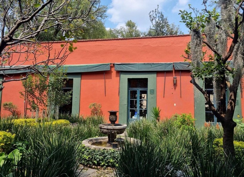 A red building with a green door, surrounded by lush greenery and a stone fountain. 2