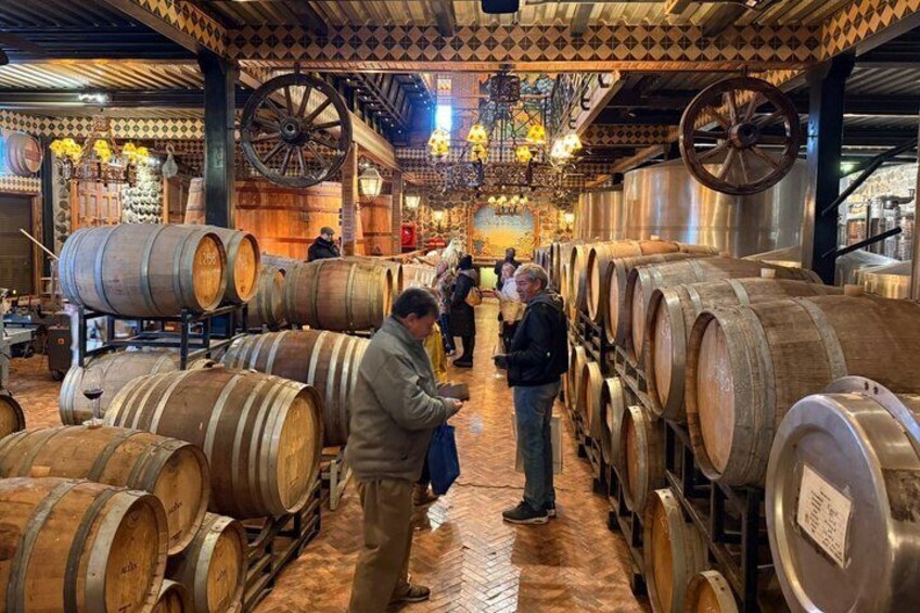 A winery with wooden and metal wine barrels, a patterned ceiling, and a central chandelier. 4