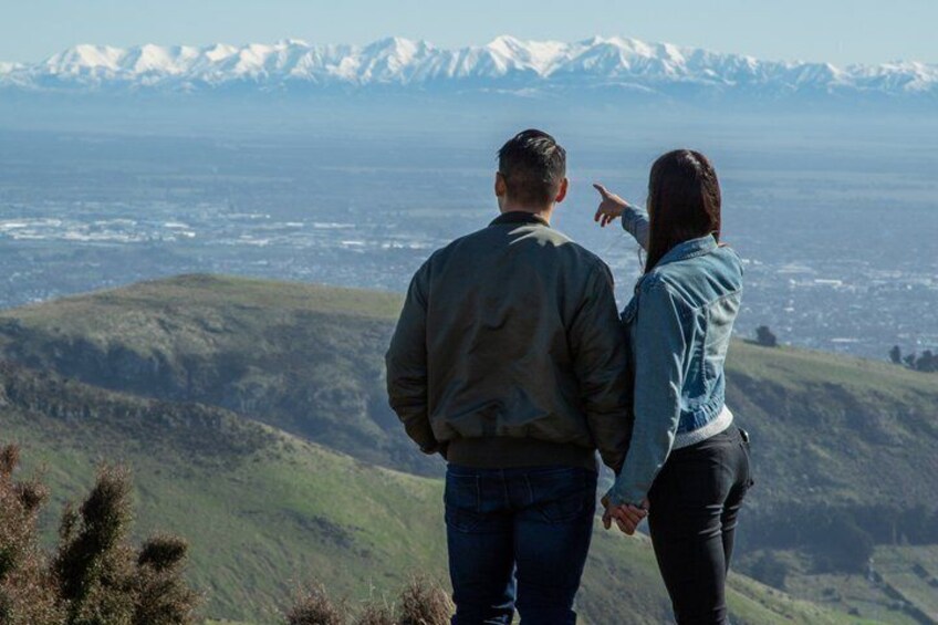 Two people standing on a hill overlooking a valley with snow-capped mountains in the distance. 1