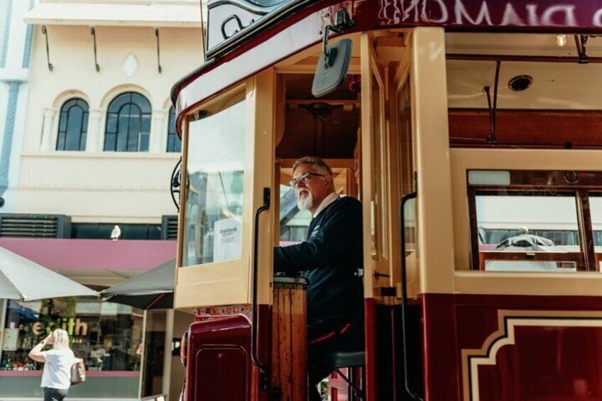 A vintage tram with a man standing at the door, a building with arched windows in the background, and a person walking on the street. 3