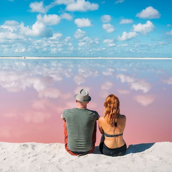 Two people sitting on a beach, facing a calm sea with a pinkish hue and a clear sky with scattered clouds. 1