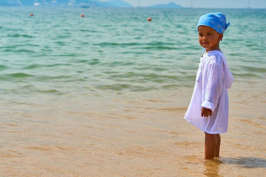 A child standing in shallow water at a beach with a blue hat and a white shirt. 4