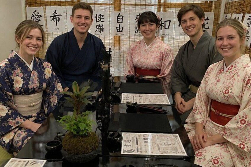 Four people in traditional Japanese attire seated at a table with a bonsai plant and menus. 4