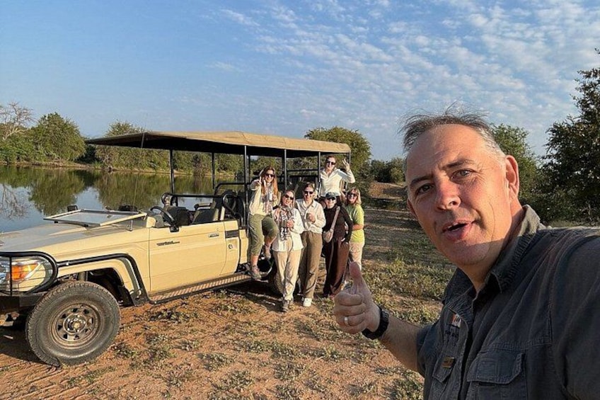 A group of people standing next to a safari vehicle on a dirt path with a body of water and trees in the background. 1