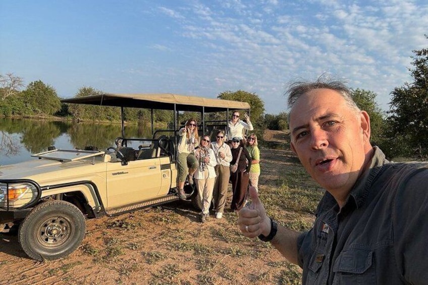A group of people standing next to a safari vehicle by a lake. 1