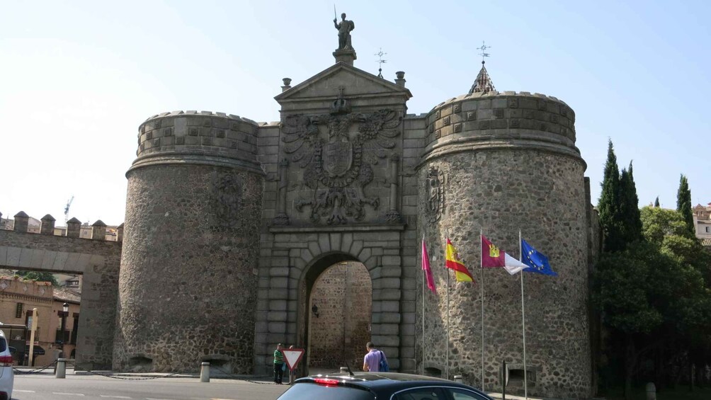 A historic stone gate with a coat of arms, flanked by two cylindrical towers, and flags of Spain and the European Union. 3