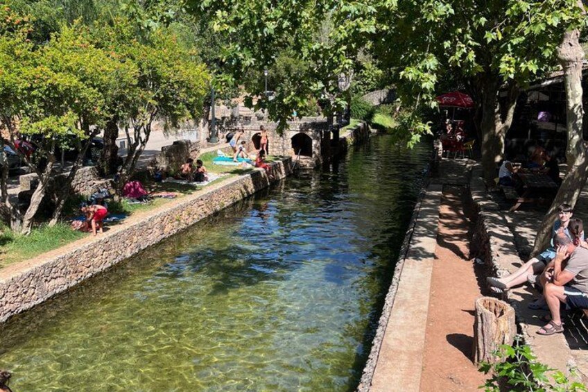 A narrow waterway with people relaxing by the banks, trees lining the path, and a stone bridge in the background. 2