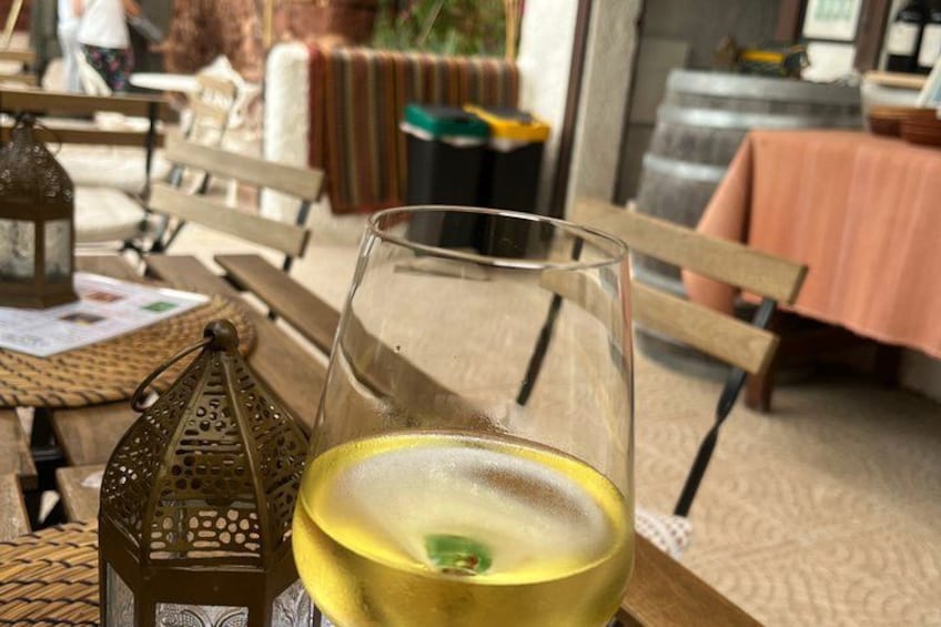 A glass of white wine on a wooden table with a woven placemat, a lantern, and a bench in a cozy outdoor setting. 5