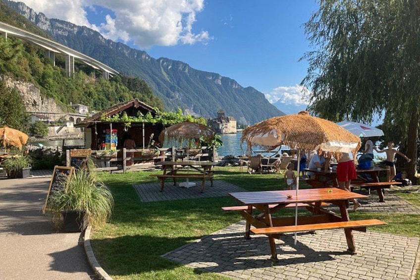 A lakeside outdoor seating area with tables and umbrellas, surrounded by mountains and greenery. 3