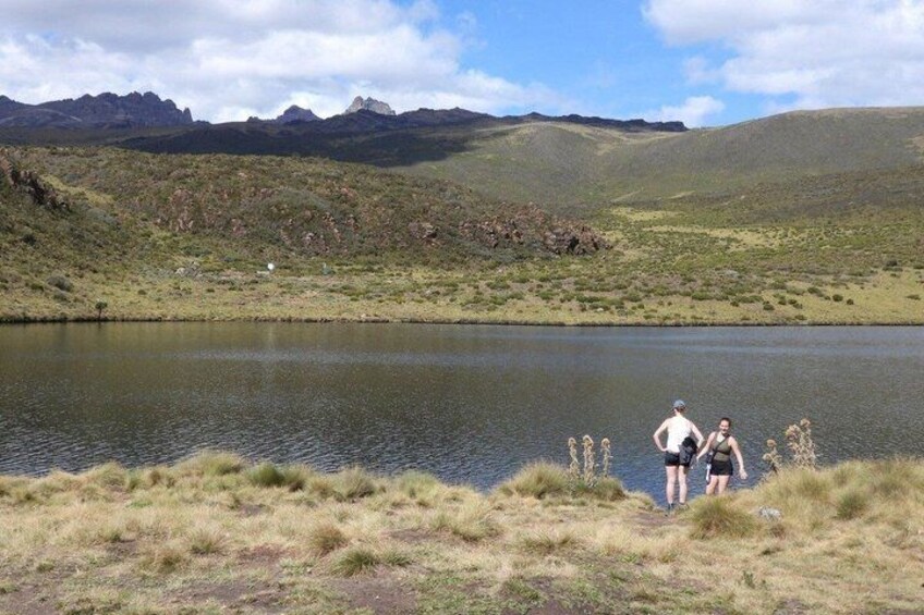A lake with grassy banks, a person standing by the water, and mountains in the background. 2