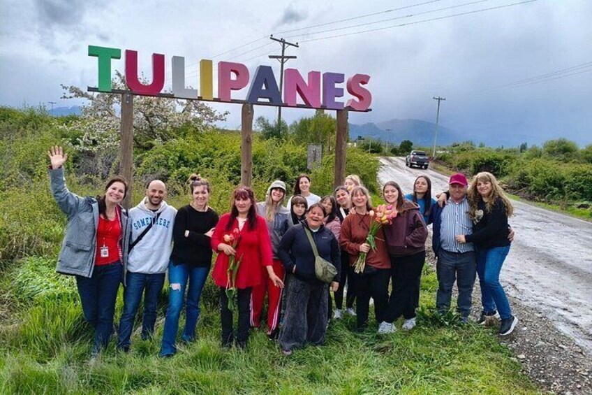 A group of people standing in front of a sign that reads 'TULIPANES' with a road and mountains in the background. 2