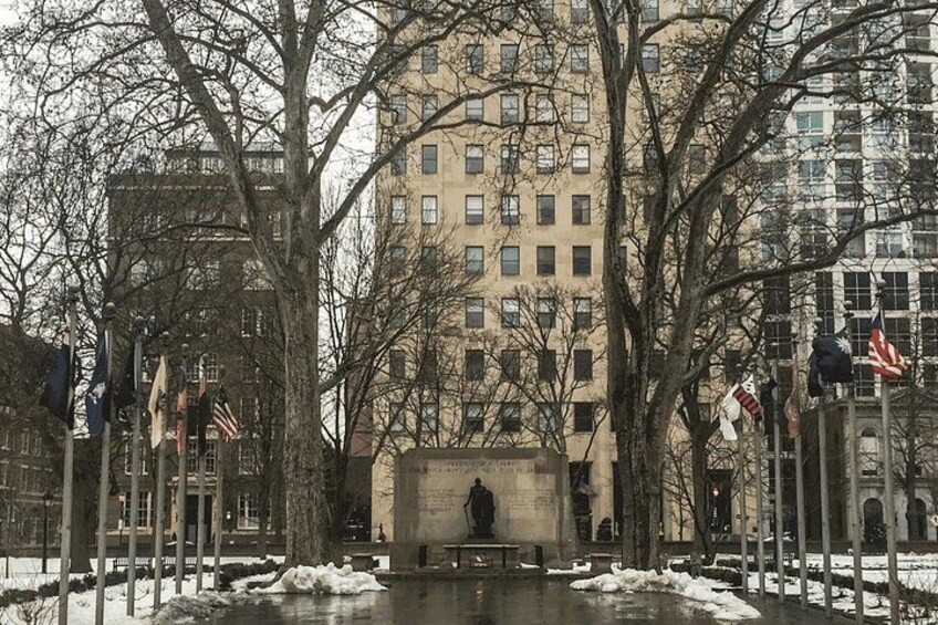 A park with a statue, trees, and a building in the background. 2