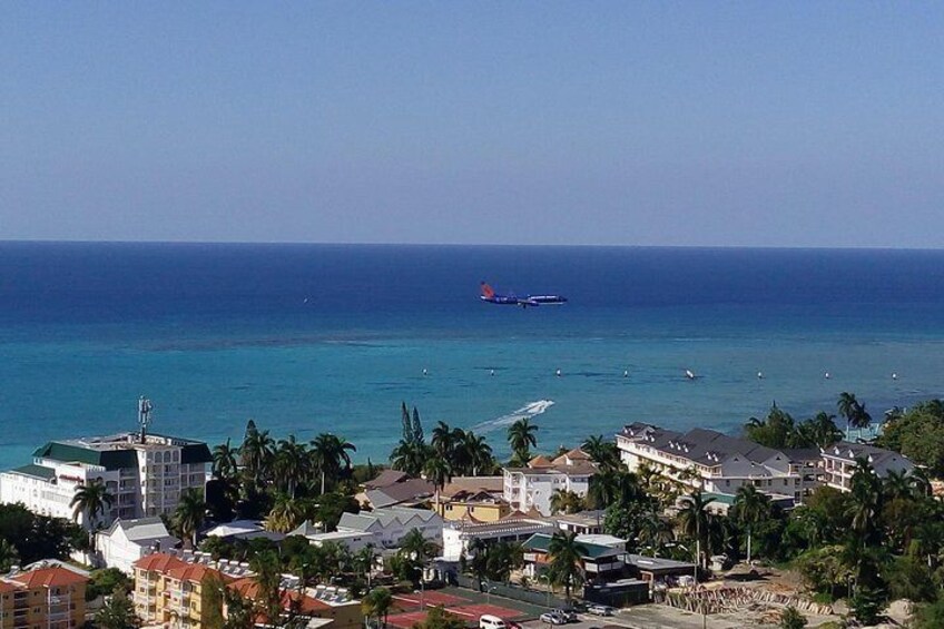 A coastal town with buildings, palm trees, and a clear blue sea. 1