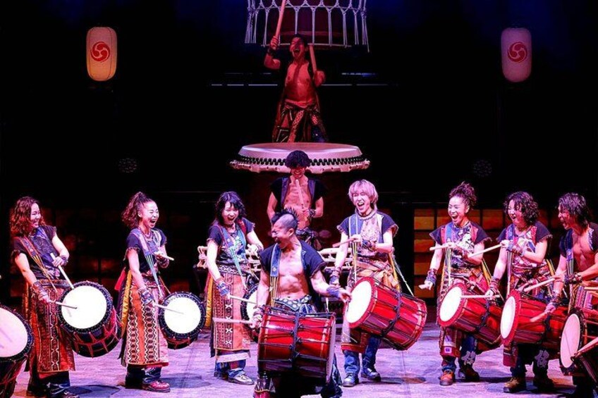 A group of performers on stage with drums, a raised platform, and a spotlight. 3