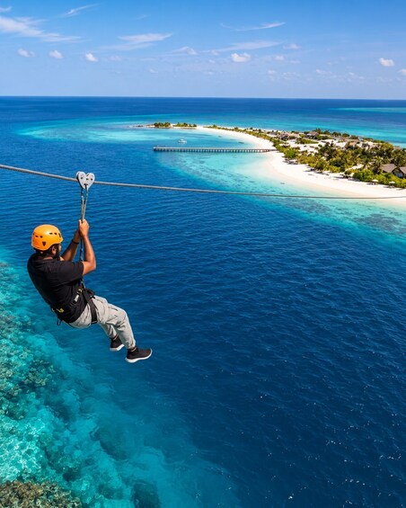A person zip-lining over turquoise waters with a beach and palm trees in the background. 1