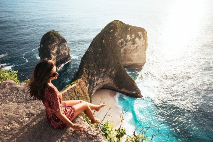 A person sitting on a cliff overlooking a coastal area with large rock formations and a beach. 2