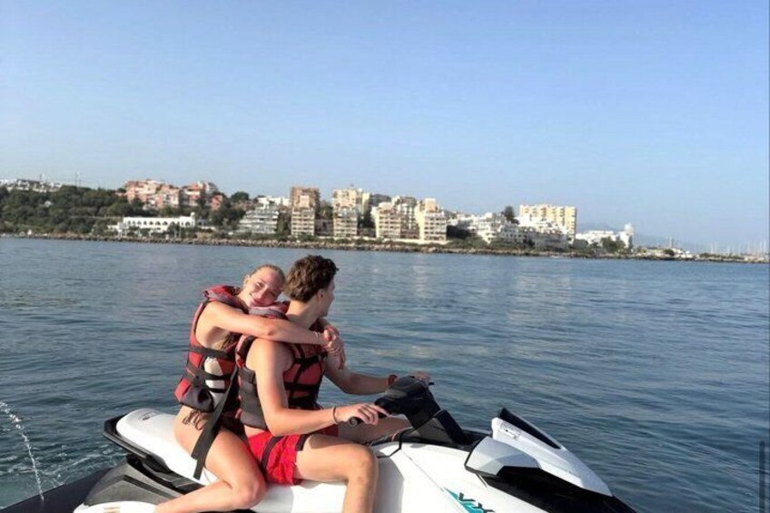 A person on a jet ski with another person on their back, water in the background, and buildings on the shore. 1