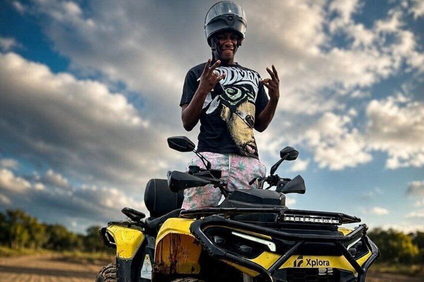 A person on a yellow ATV with a helmet, standing on a dirt path with trees and a cloudy sky in the background. 2