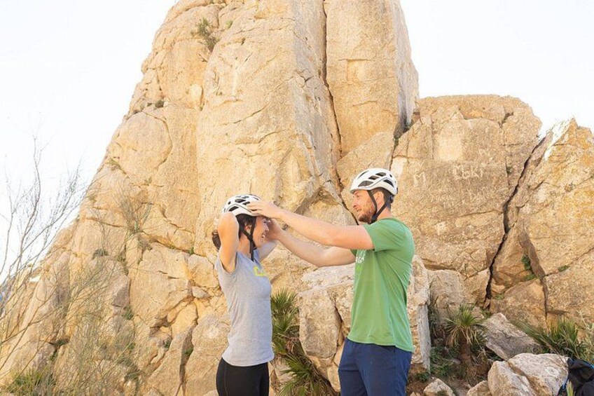 Two people wearing helmets, one in a green shirt and the other in a grey shirt, standing in front of a large rock formation. 3