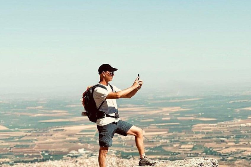 A person taking a selfie with a smartphone, wearing a cap, shorts, and a backpack, standing on a rocky outcrop with a vast landscape in the background. 2