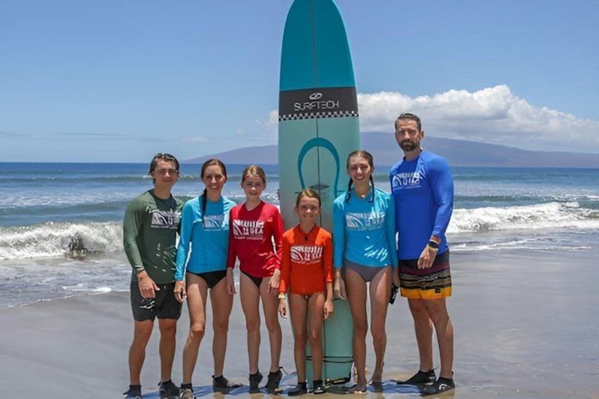 A group of people standing on a beach with a surfboard in the background. 1