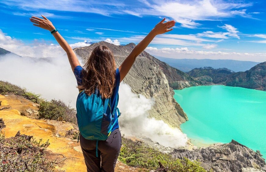 A person standing on a mountain with arms raised, overlooking a turquoise lake and a volcanic landscape. 4