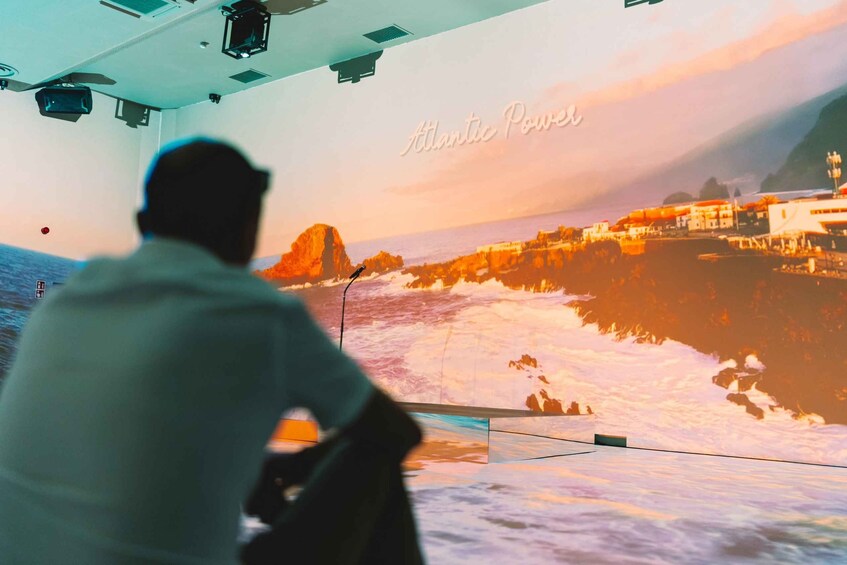 A person in a white shirt looking out at a coastal scene with waves crashing onto rocks and buildings in the background. 3
