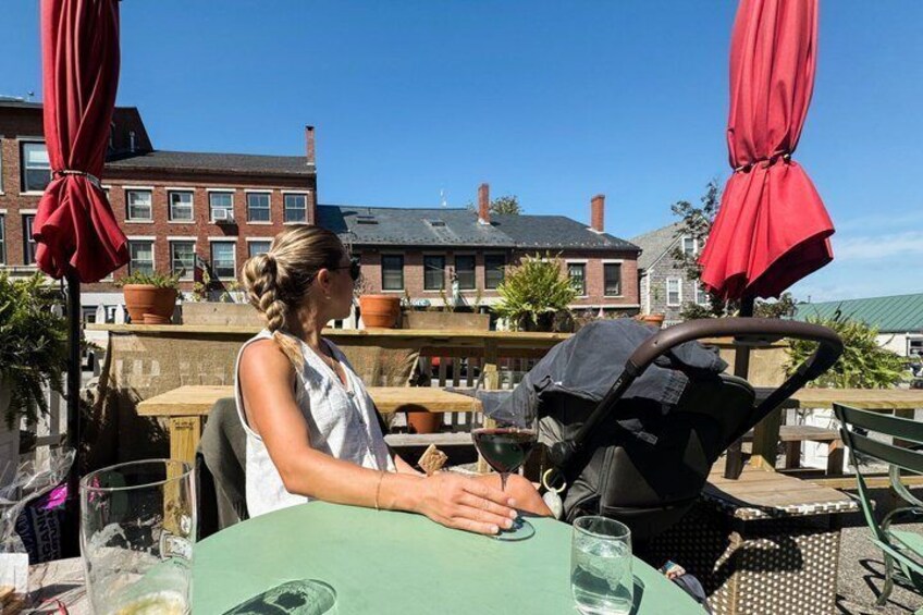 A person seated at an outdoor table with a stroller, red umbrellas, and buildings in the background. 3