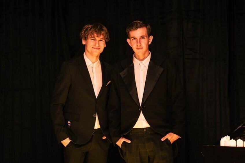 Two men in black suits with white shirts standing in front of a black curtain. 4