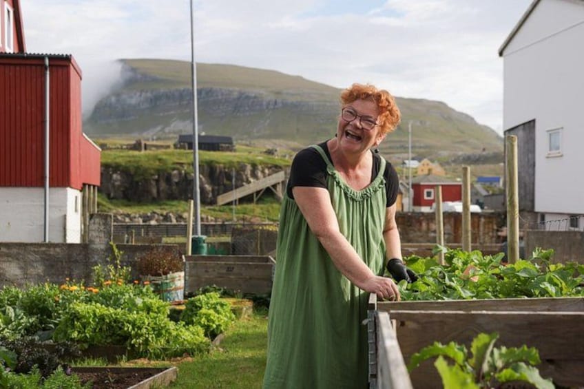 A person tending to a garden with vegetables, a red building, and a mountainous landscape in the background. 2